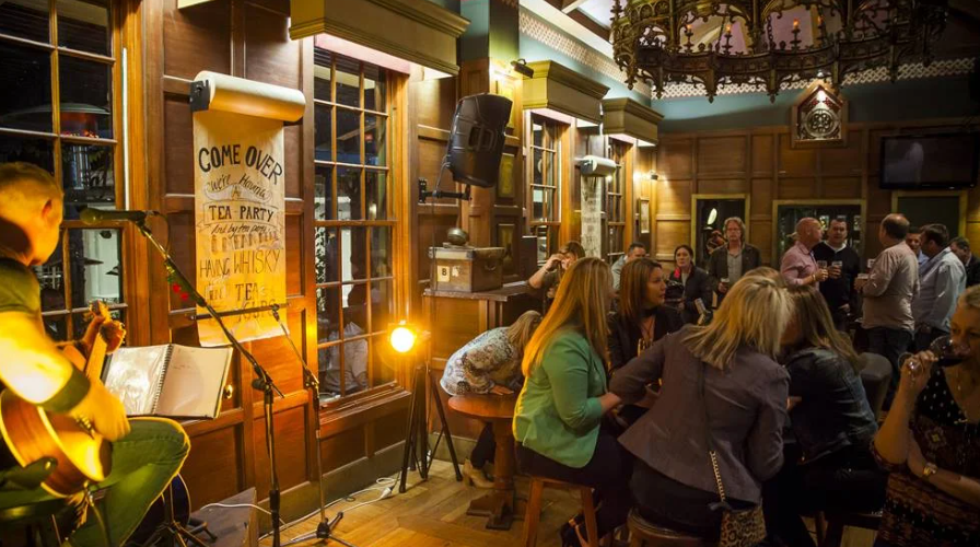 The Fiddler pub interior with warm timber panelling, live acoustic music, patrons enjoying drinks and atmospheric chandelier lighting
